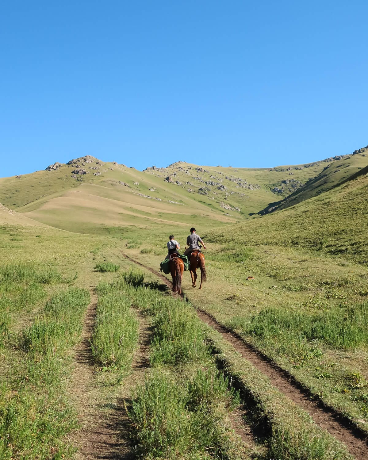 Ari and our guide on horseback riding along a dirt track through a valley surrounded by green hills.