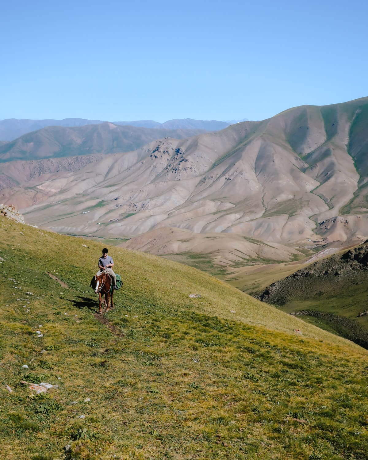 Ari on horseback following a narrow dirt path through green mountain slopes with rolling peaks in the distance.