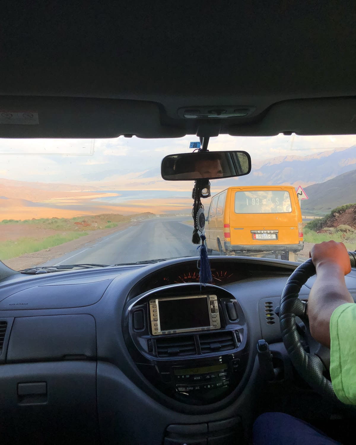 View from inside a car driving on a winding road, passing a yellow van with distant mountains in the background.