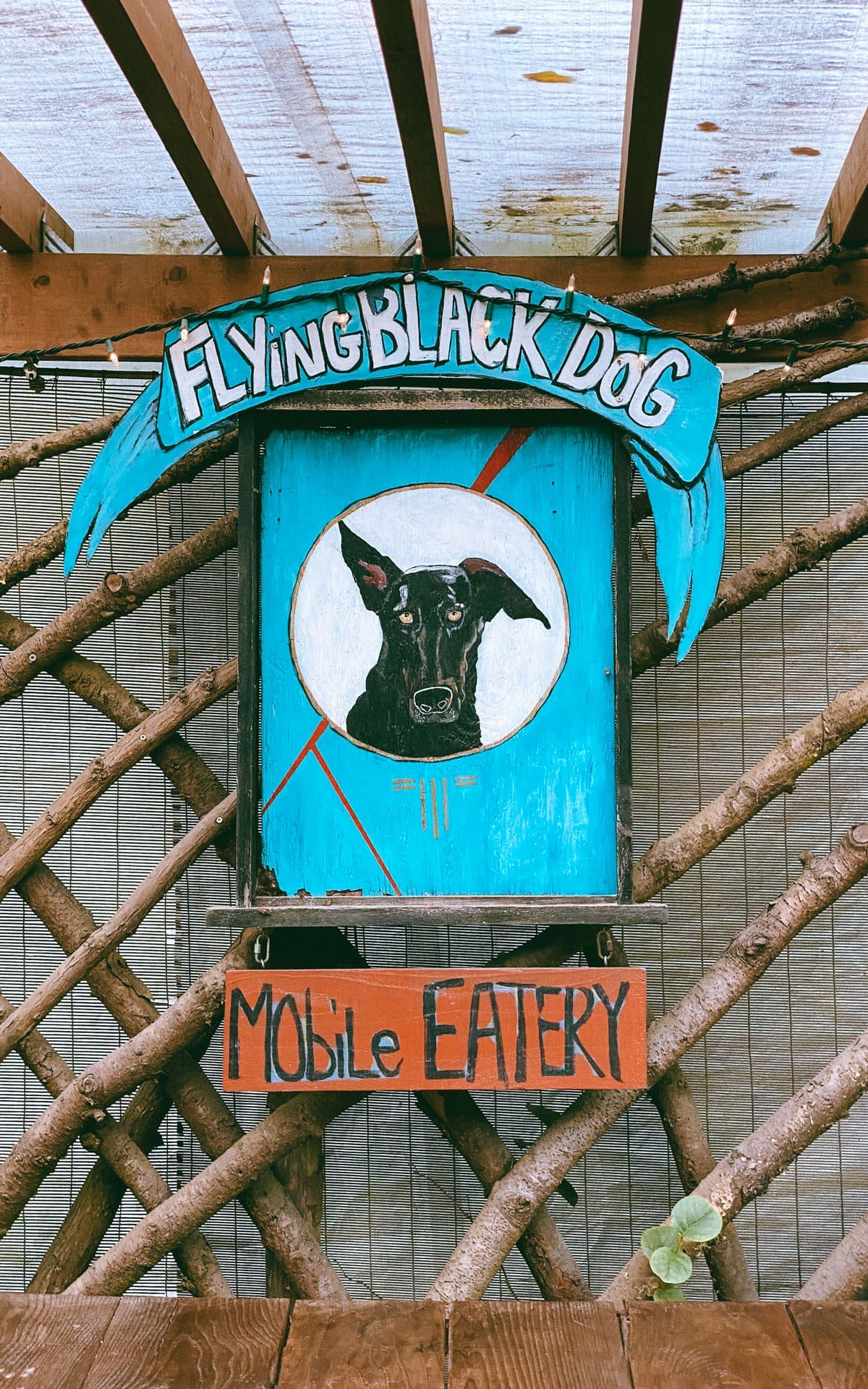 A hand-painted wooden sign for Flying Black Dog Mobile Eatery, featuring a black dog illustration on a turquoise background