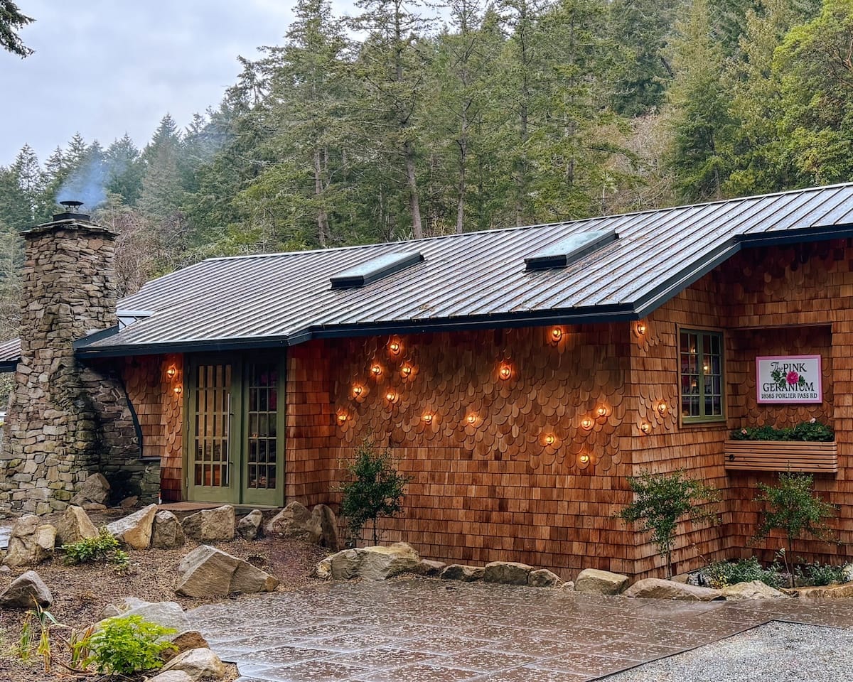 A cozy cedar-shingled restaurant with glowing lights, a stone chimney, and forest surroundings at The Pink Geranium, Galiano Island