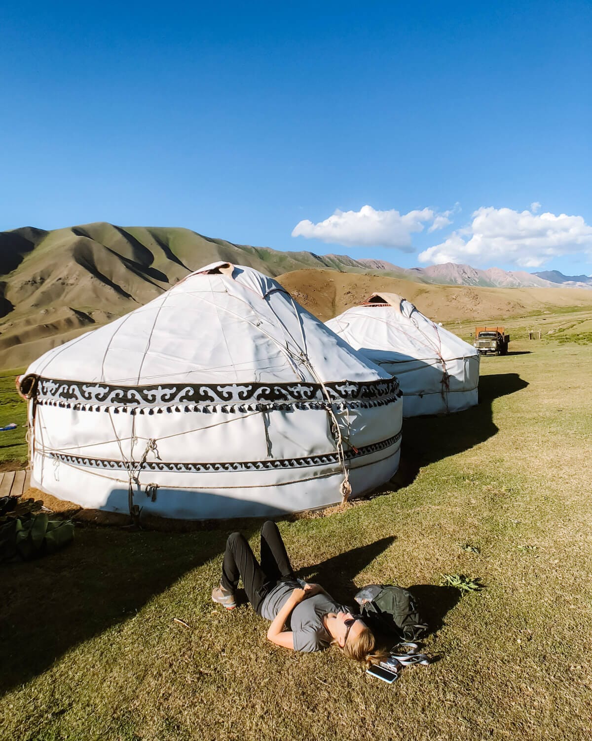 Cec lying on the grass beside a row of white yurts with decorative trim, surrounded by green hills.