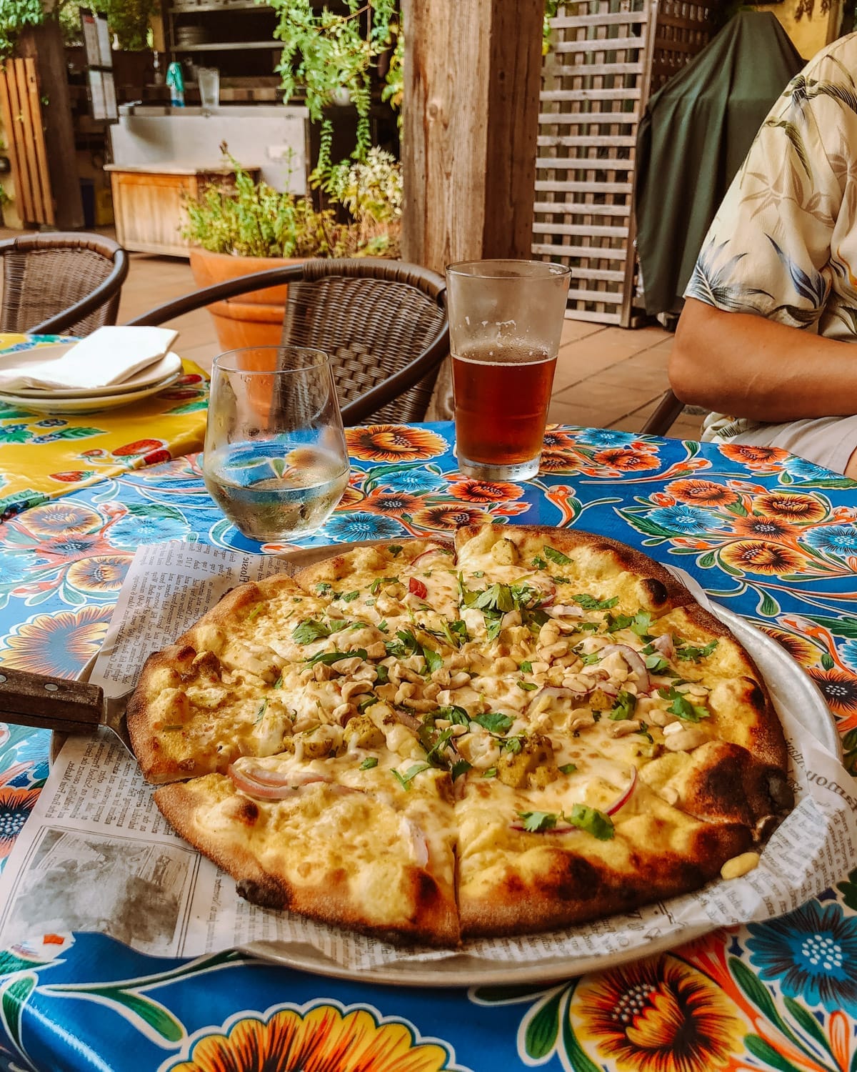 A wood-fired pizza with cheese, cashews, and herbs on a newspaper-lined plate, set on a colourful outdoor table at La Lena Pizza Patio.