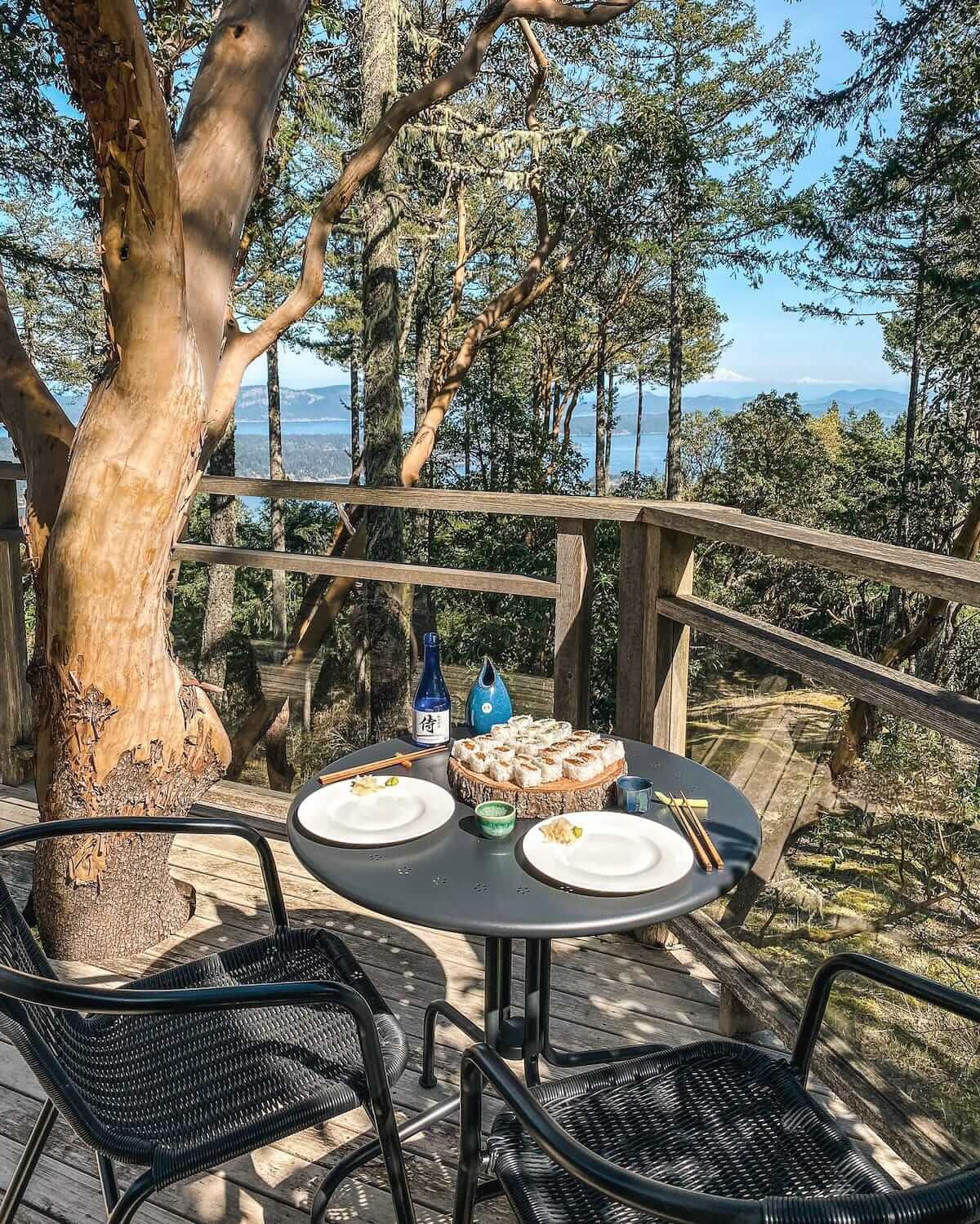 Two-seat outdoor dining setup with sushi and sake on a wooden deck overlooking forest views on Salt Spring Island.