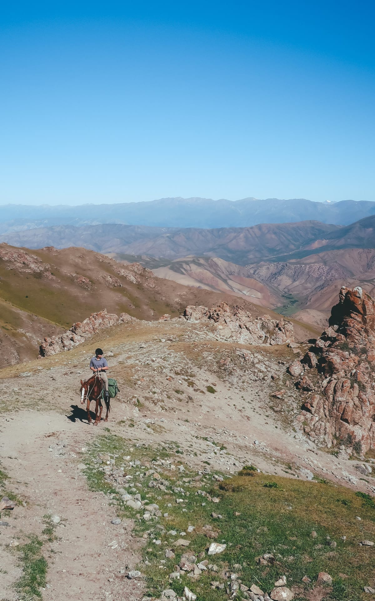 Ari descending a rugged mountain trail on a horse towards Song Kul Lake, surrounded by valleys and distant peaks.