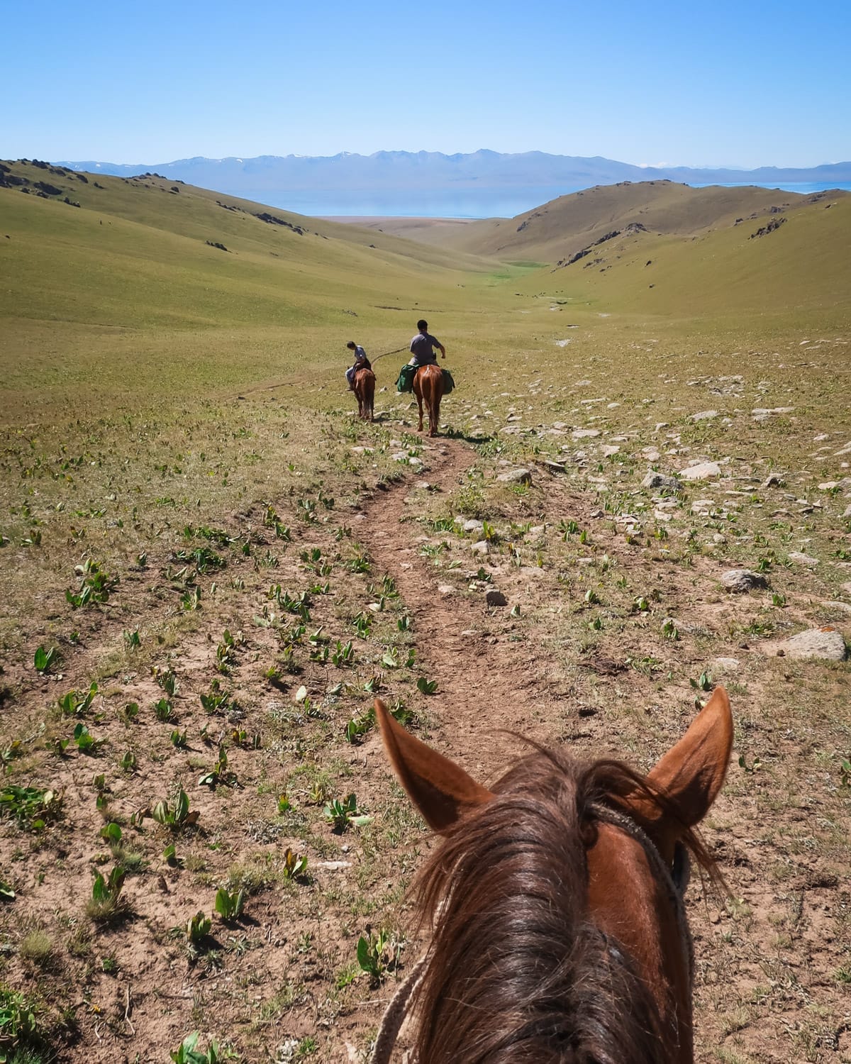 Two horseback riders taking on a scenic dirt trail in a Kyrgyz valley, with distant views of mountains and Song-Kul Lake.