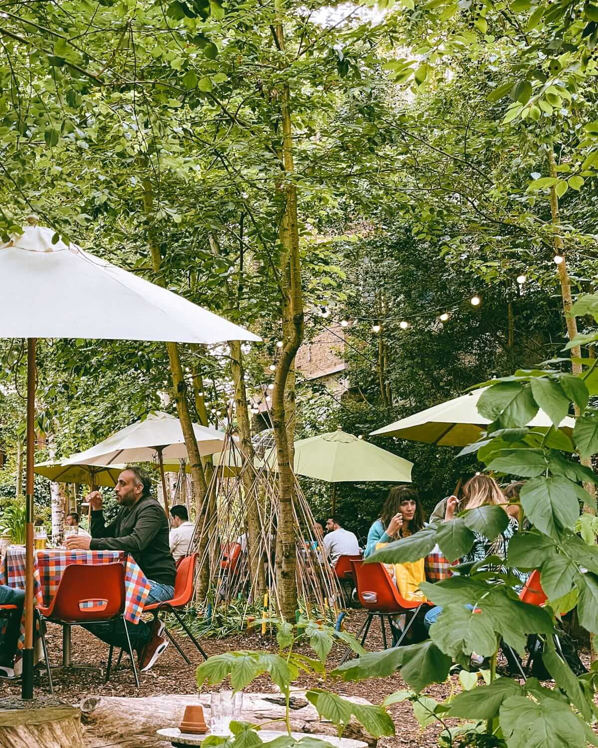 People dining under umbrellas in the lush greenery of Dalston Curve Garden, with fairy lights adding charm to the outdoor seating.