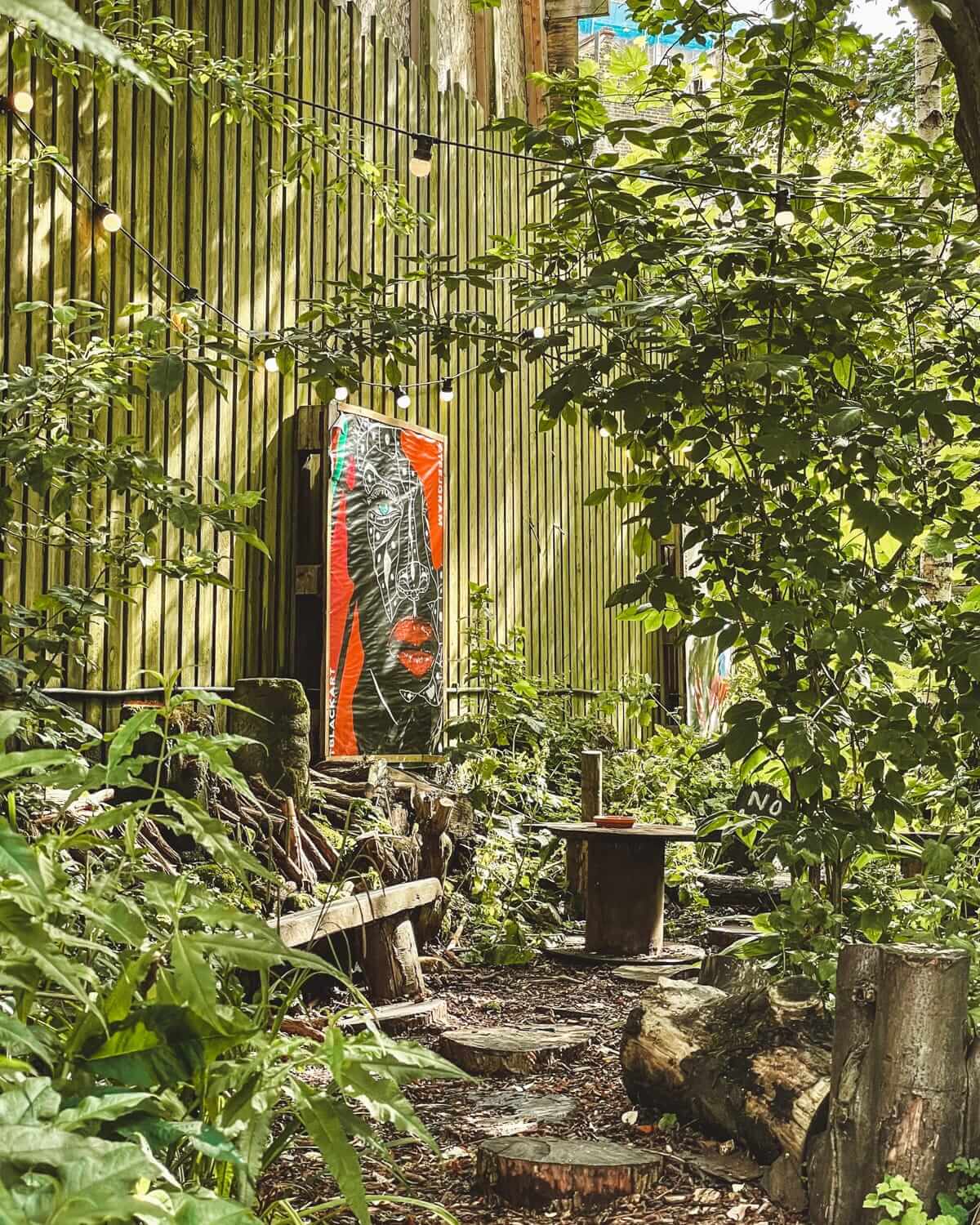 A hidden seating area in Dalston Curve Garden, surrounded by foliage, with a contemporary piece of artwork and string lights.