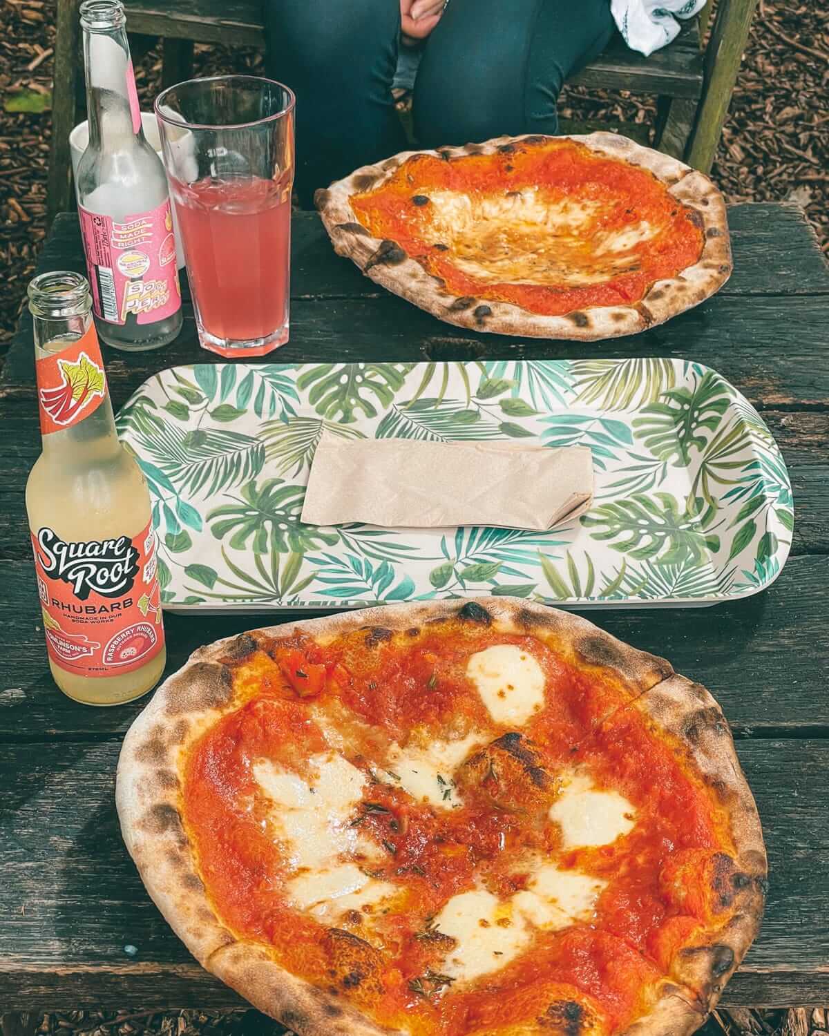 Two margarita pizzas and rhubarb sodas on a wooden table outside.