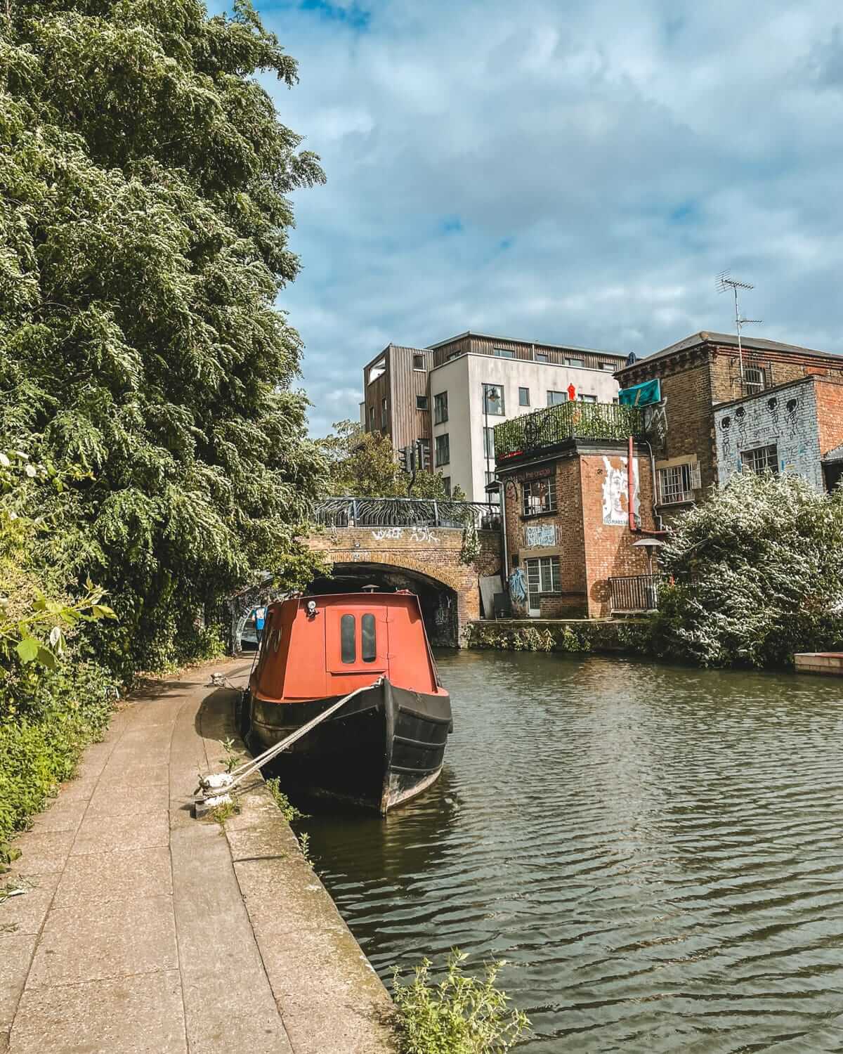 A red houseboat moored along Regent’s Canal, with a tree-lined path leading to a graffiti-adorned brick bridge in East London.