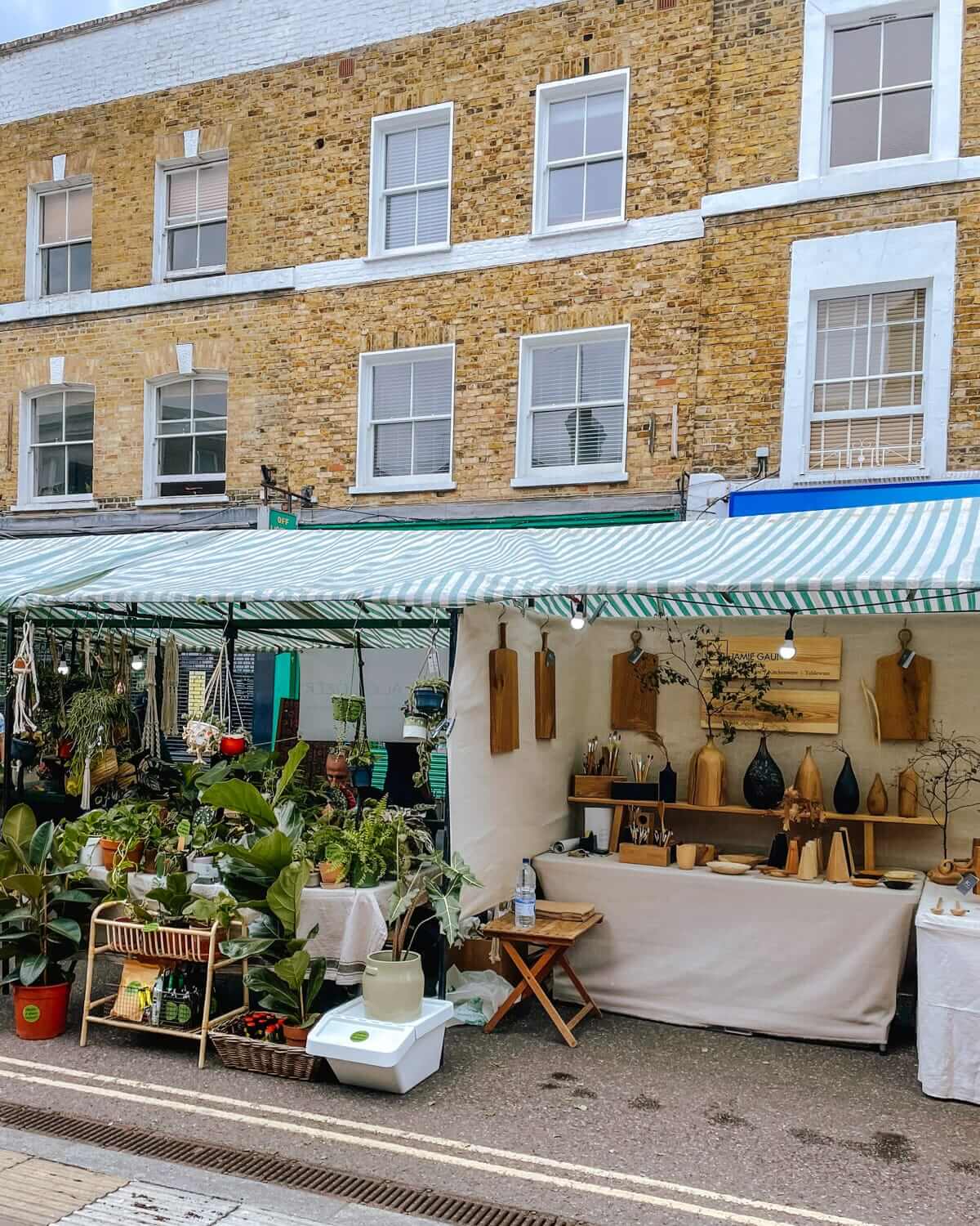 A street market stall with plants, wooden crafts, and home decor under a green and white striped canopy at Broadway Market.