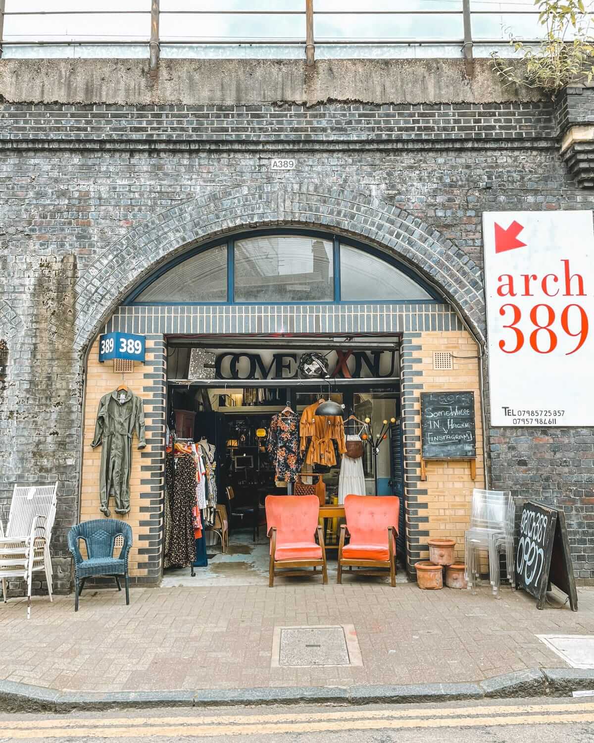 A vintage store inside a brick railway arch, with clothing, decor, and two retro chairs outside.