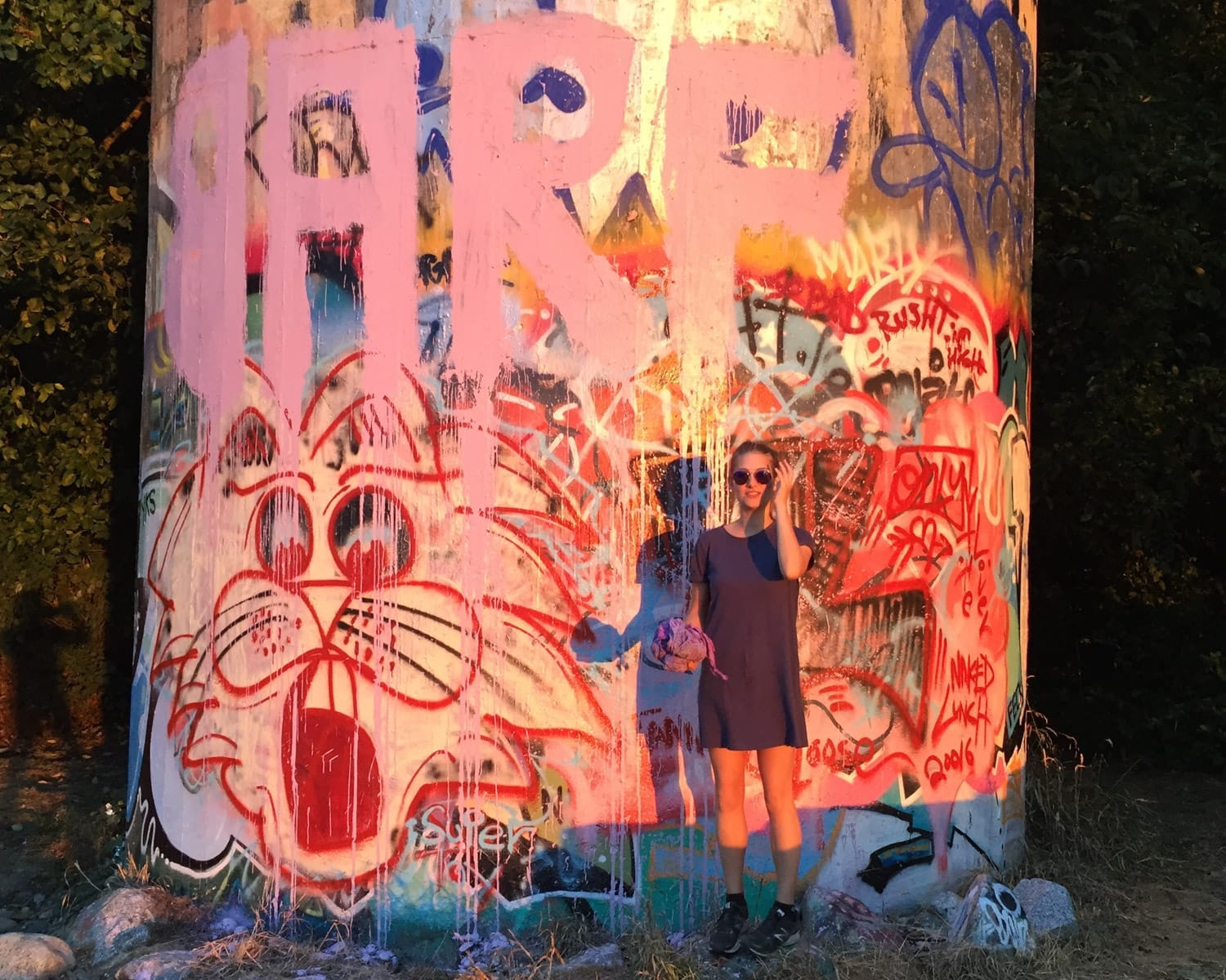 Cec in a t-shirt dress standing by a concrete structure with vibrant street art on the coast under warm evening sunlight.