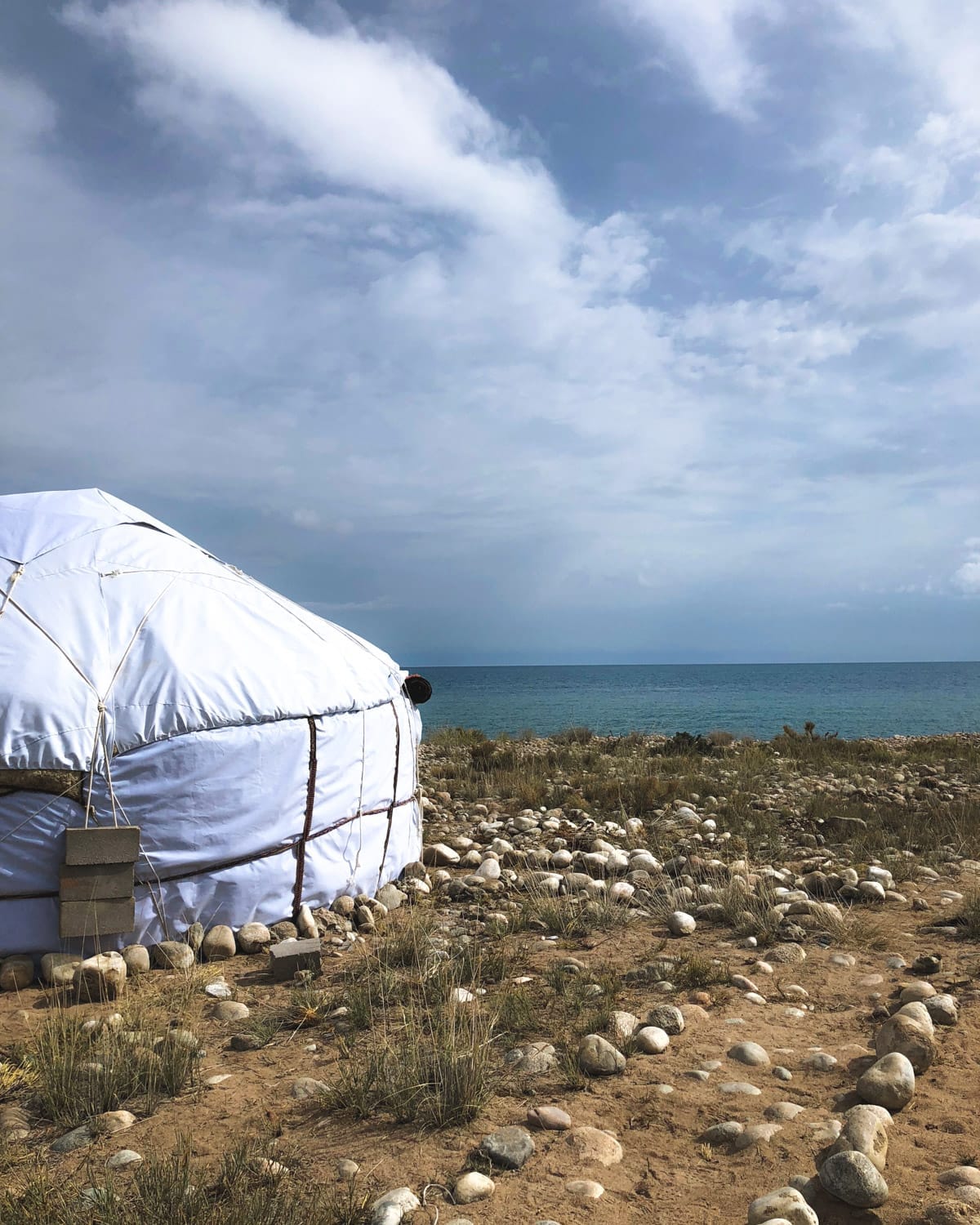 White yurt set on a rocky shoreline of Issyk-Kul Lake, with blue waters and cloudy skies in the background.