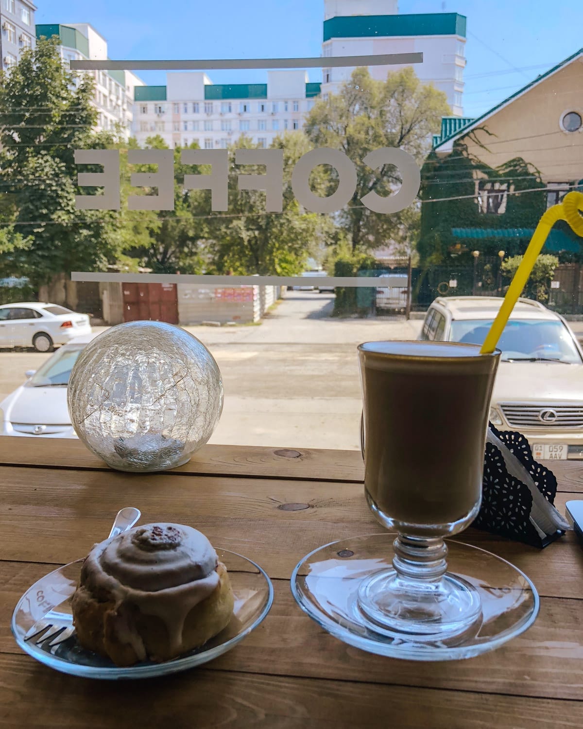  Latte and a cinnamon roll on a wooden table at a cafe in Bishkek, Kyrgyzstan, with a view of parked cars outside.