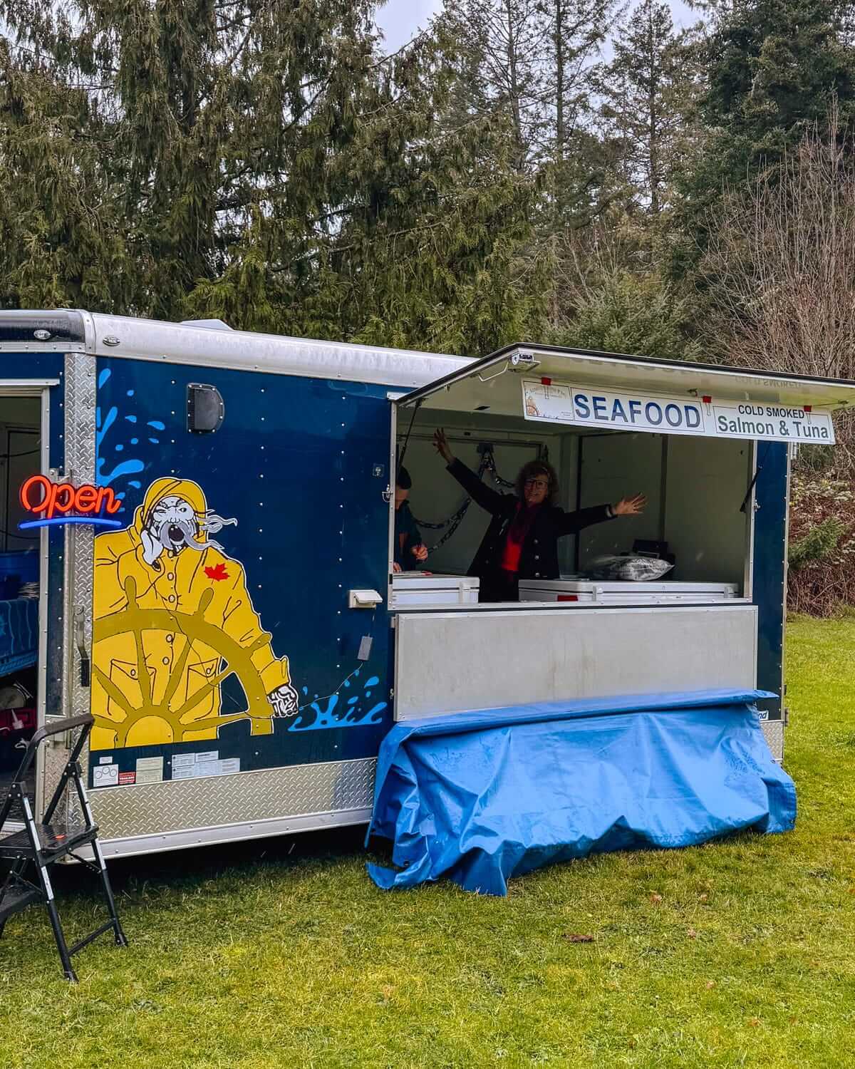 Seafood vendor in a vibrant blue food truck with a fisherman illustration in a forested setting.