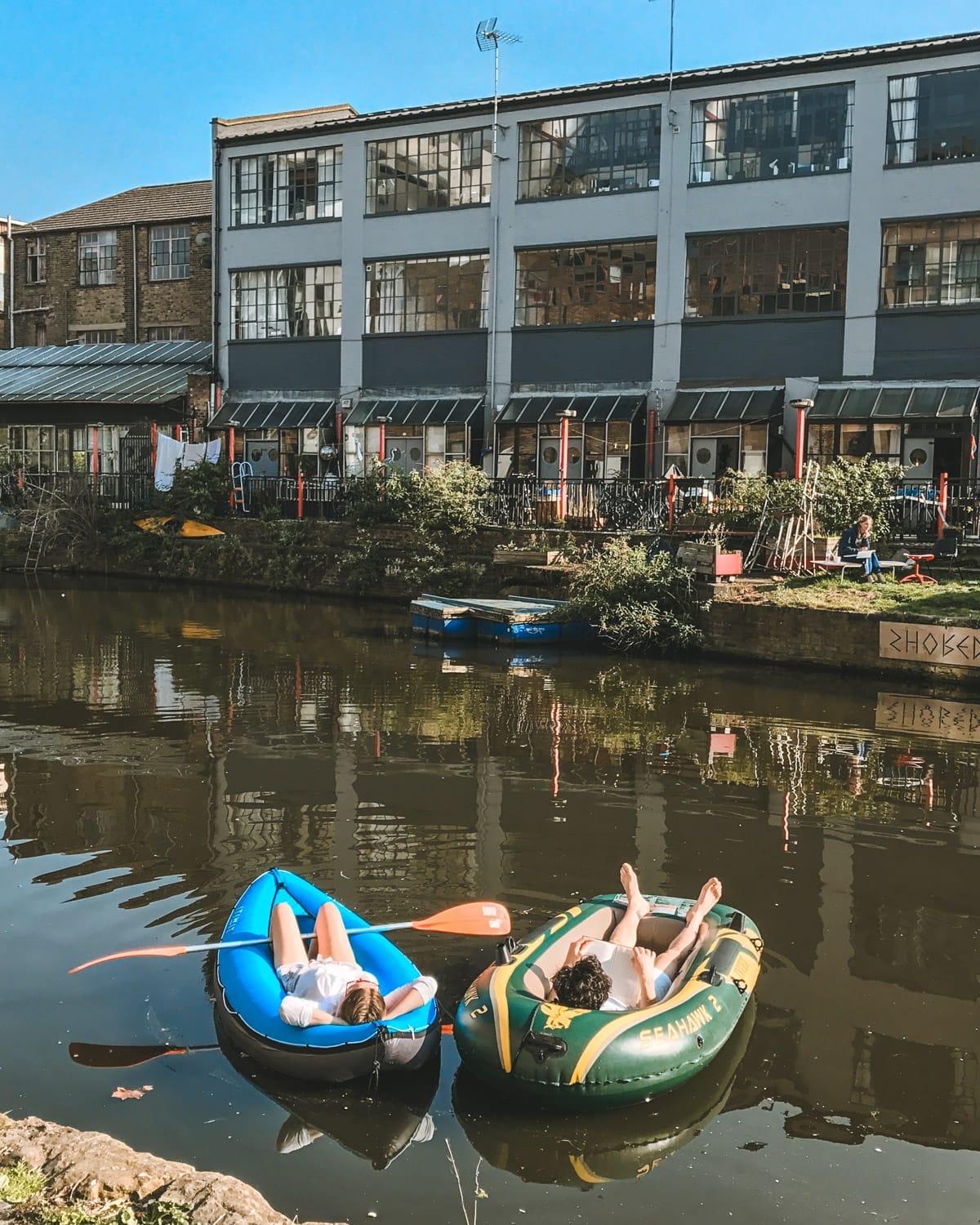 Two people lounging in inflatable boats on Regent’s Canal, basking in the sun, with warehouse-style buildings in the background.