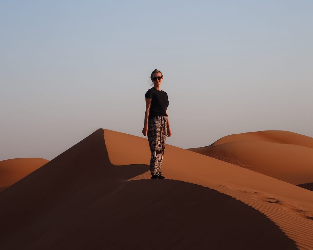 Cec standing on a sand dune in Oman under clear blue skies, wearing sunglasses, with rippled desert sand surrounding her.