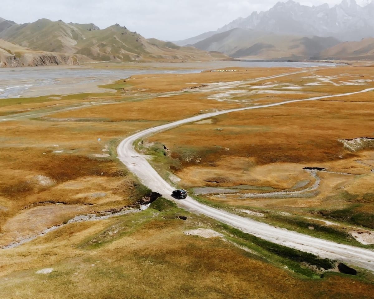 A drone shot of a dirt road winding through golden grasslands and valleys in the Tian Shan Mountains.