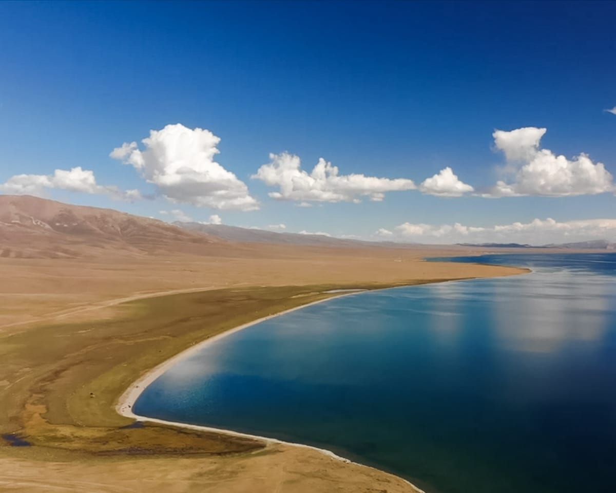 A drone shot of Song Kul Lake with calm blue water, golden grasslands and a bright sky.