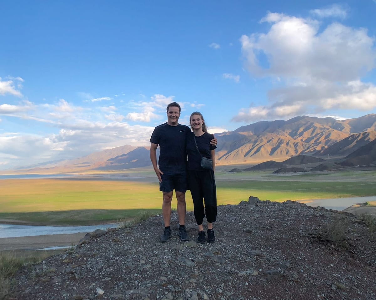 Ari and Cec standing on a hilltop in Kyrgyzstan, overlooking a field and mountains glowing from the light.