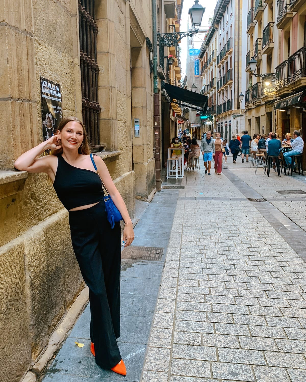 Cec posing against a wall on a cobblestone street in San Sebastián, Spain, with lively outdoor cafes nearby.