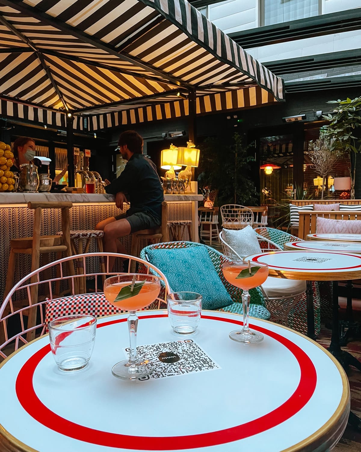 A stylish courtyard bar at Mama Shelter with black-and-white striped canopies, colourful seating, and two elegant pink cocktails.