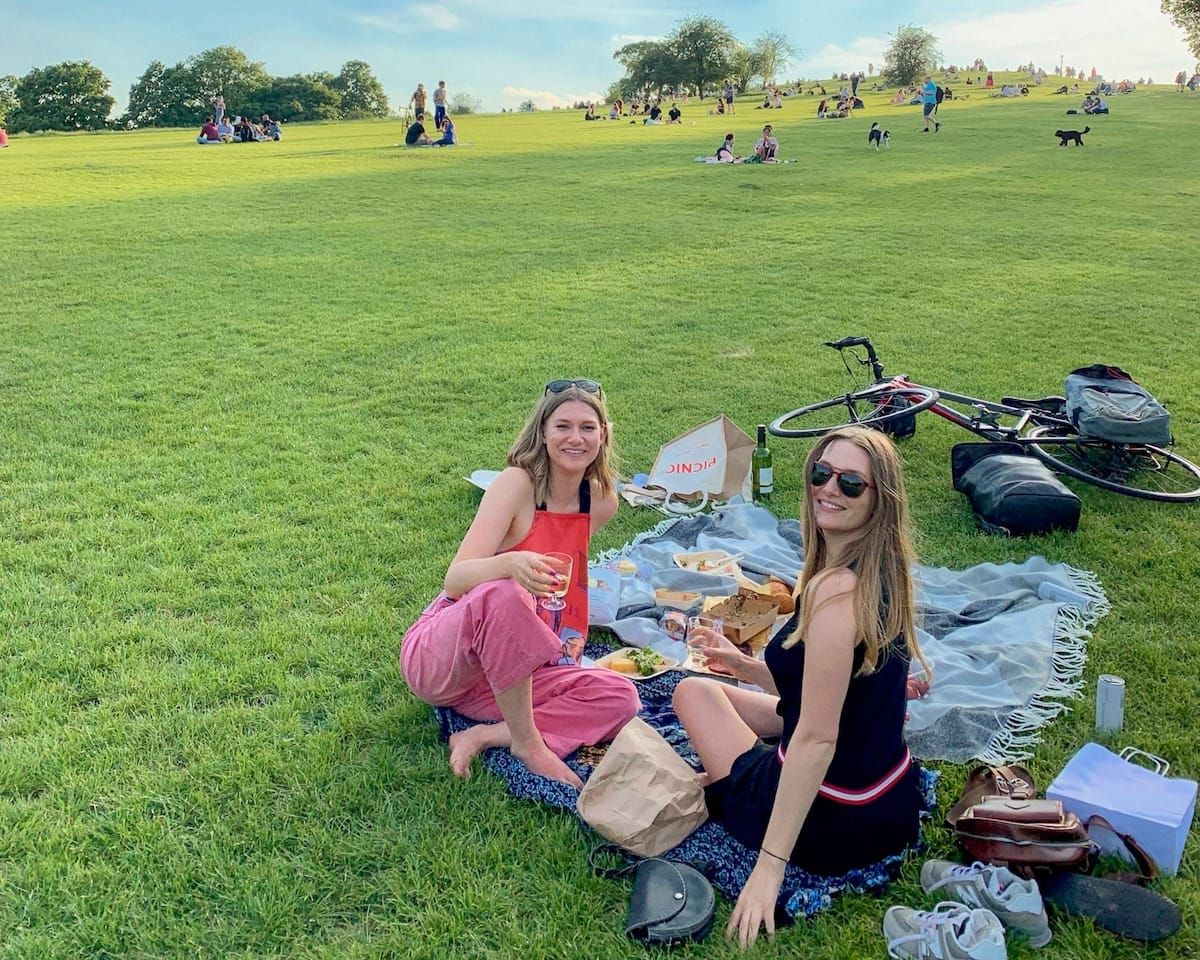 Cec and Katarina enjoying a picnic on the grassy Primose Hill, surrounded by parkgoers on a sunny day.