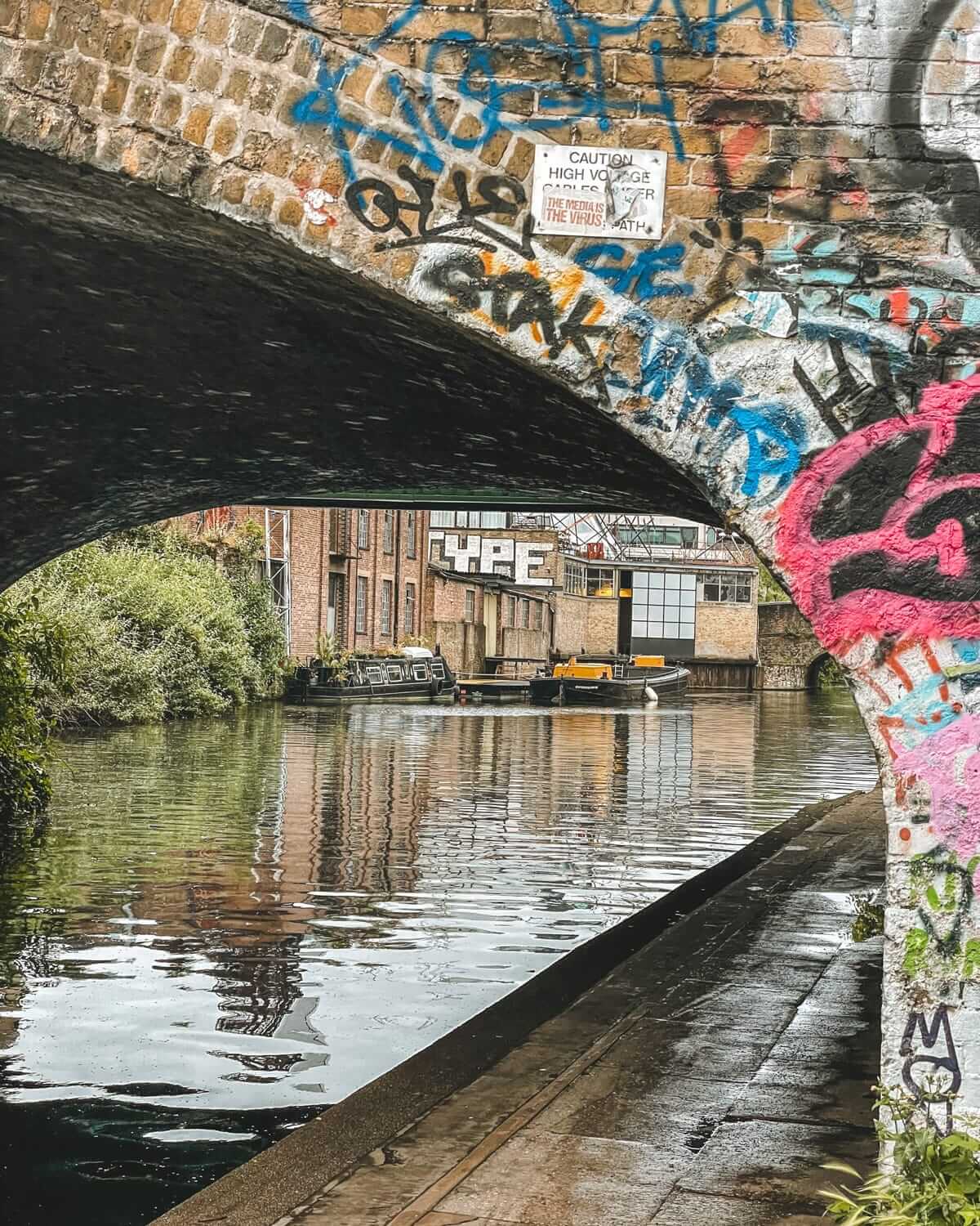 A graffiti-covered brick bridge frames a view of Regent’s Canal, with a boat, warehouse-style buildings, and greenery along the water.