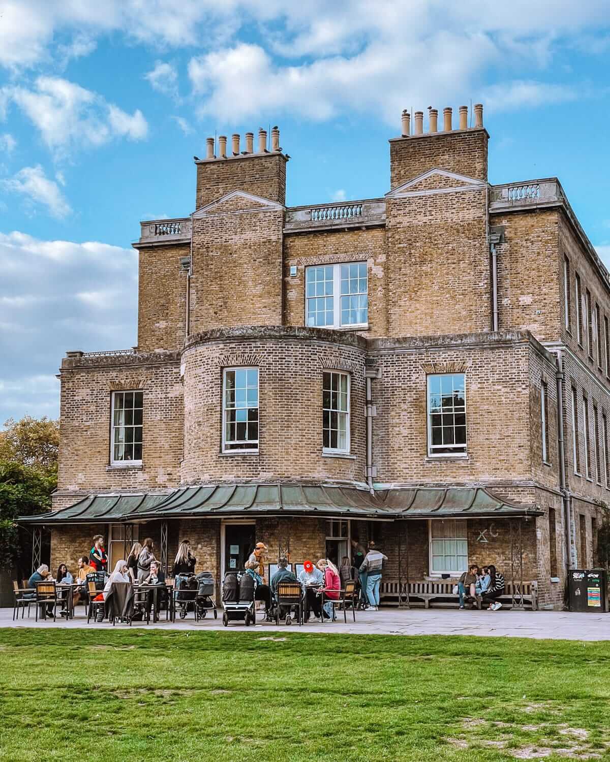 The grand brick exterior of a historic building in Clissold Park with outdoor seating where people enjoy conversation.