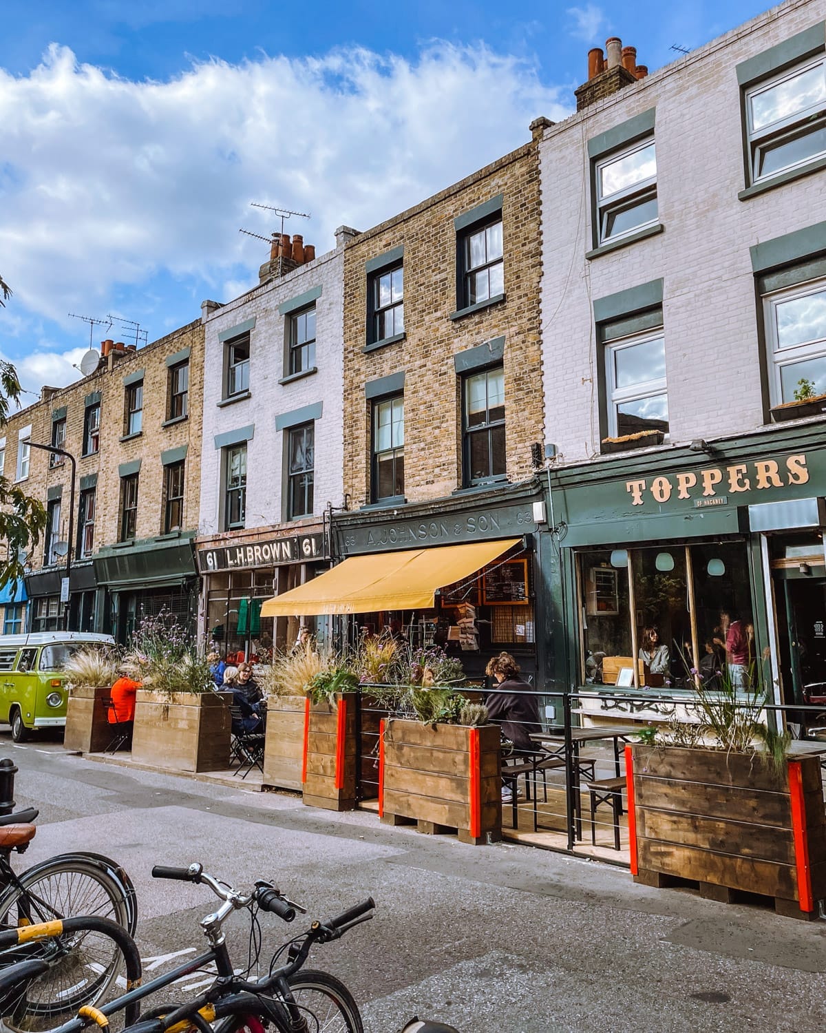 A lively street scene on Wilton Way with brick townhouses, a cafe with a yellow awning, outdoor seating and parked bicycles.