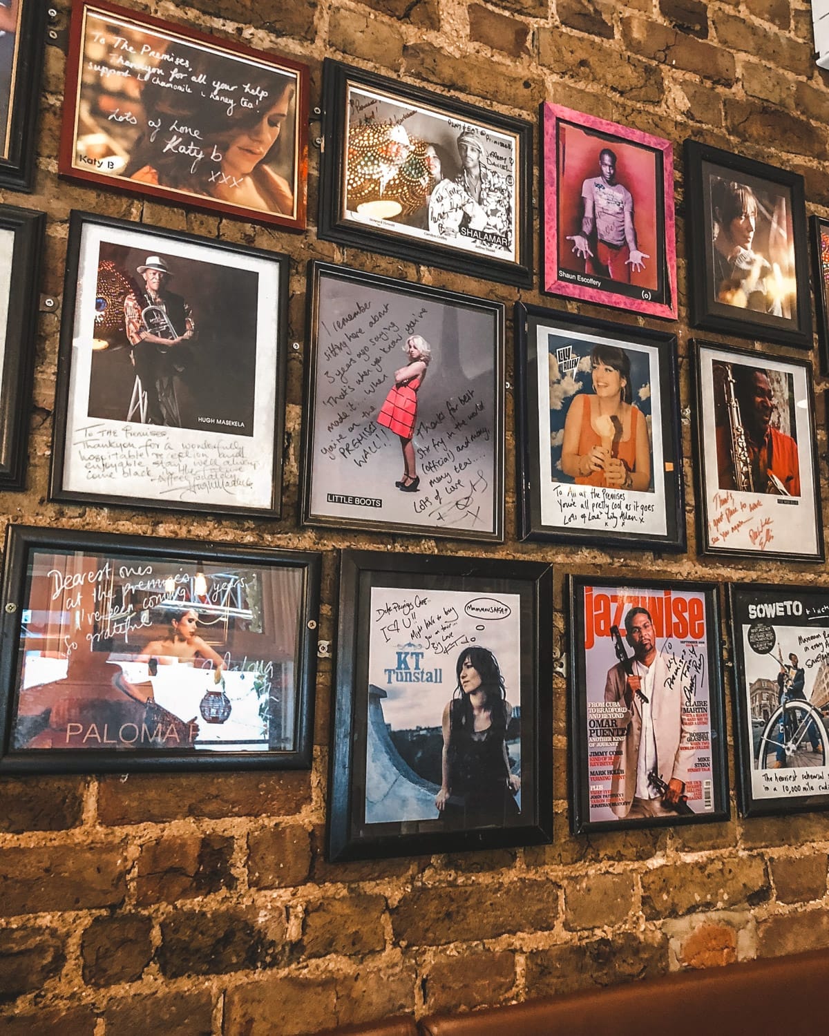 A brick wall inside The Premises Cafe adorned with framed signed photographs of musicians.