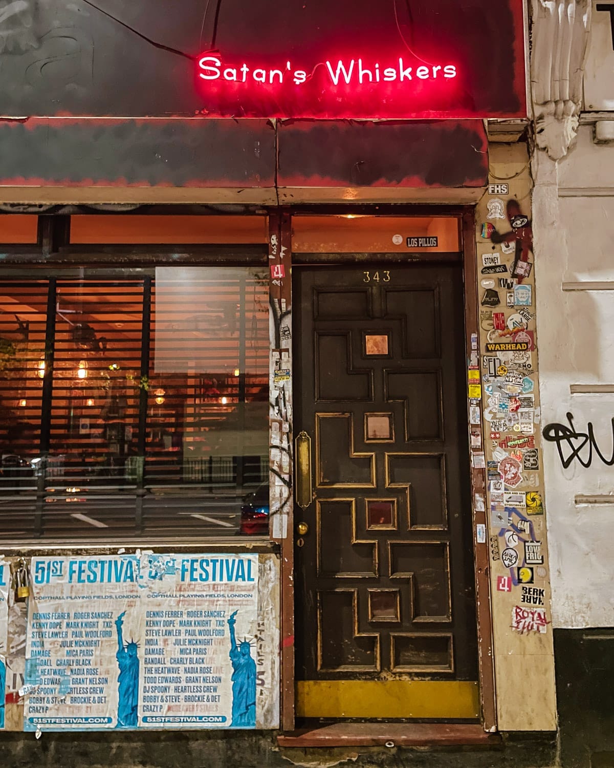 The moody bar entrance of Satan's Whiskers with a glowing red neon sign above a door covered in stickers and posters.