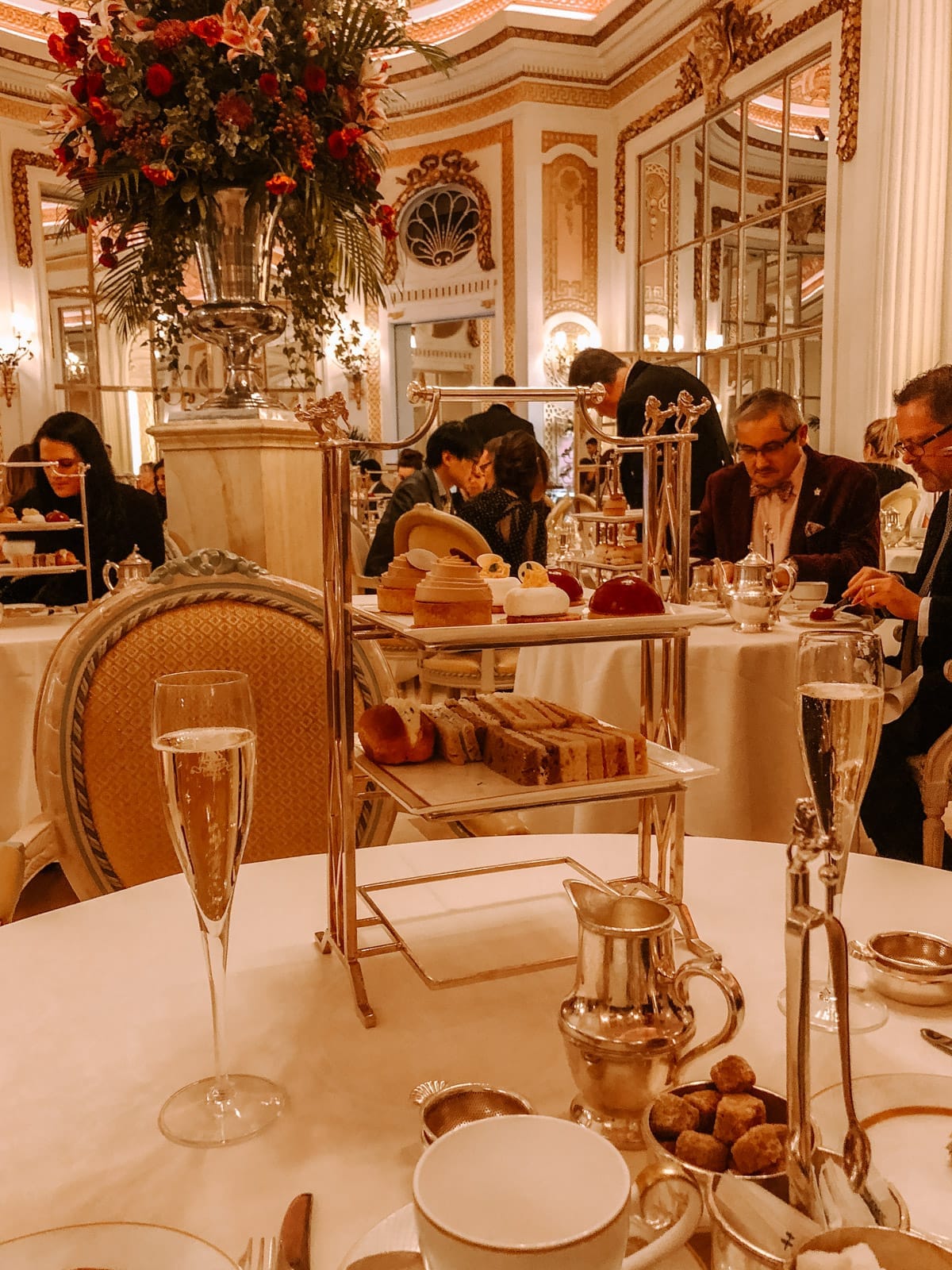 A luxurious tea setting with fine china, champagne, pastries, and finger sandwiches in an ornate dining room at The Ritz..