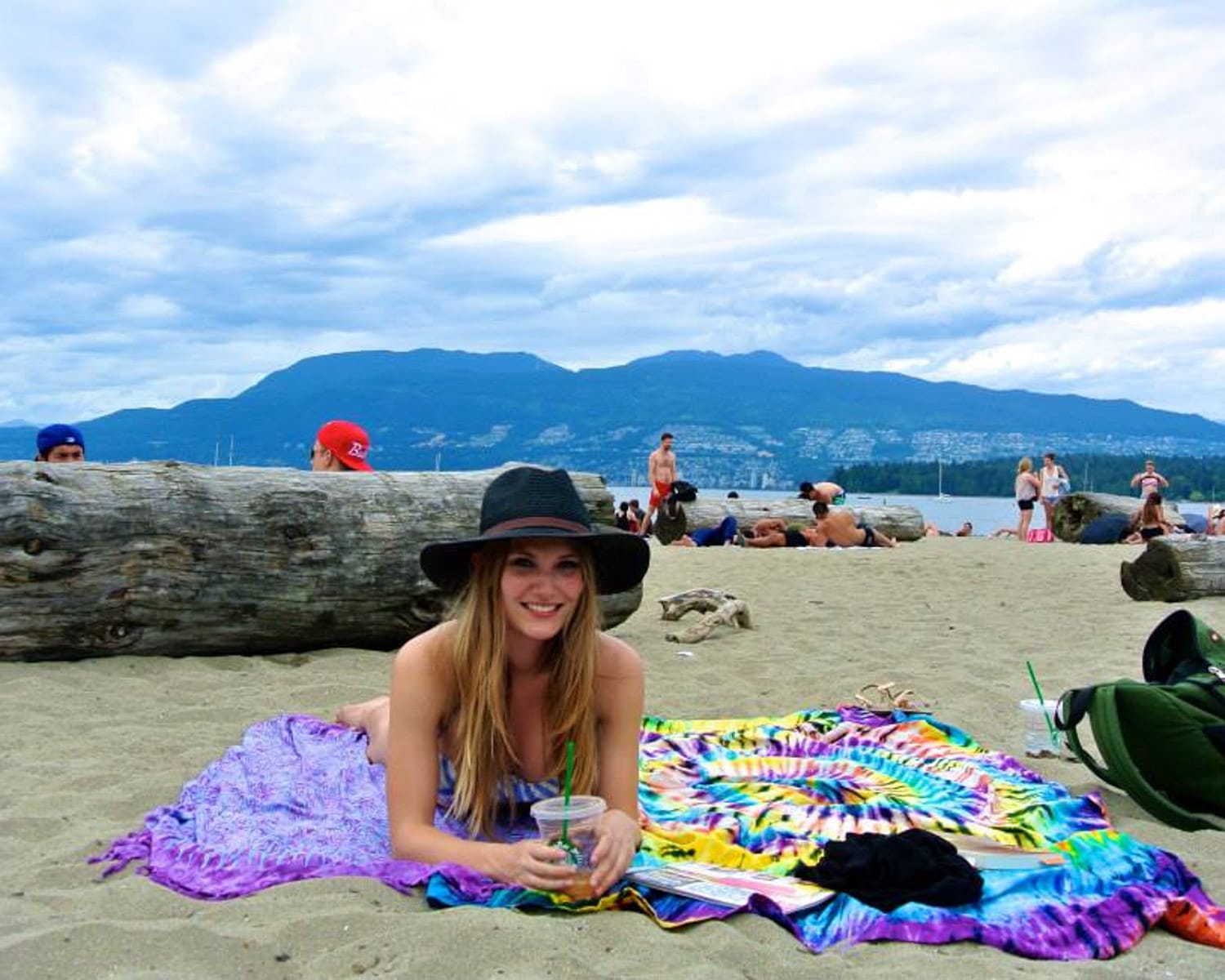 Cecily relaxing on Kits Beach in Vancouver, lying on a colourful tie-dye blanket, holding a drink with mountains in the background.