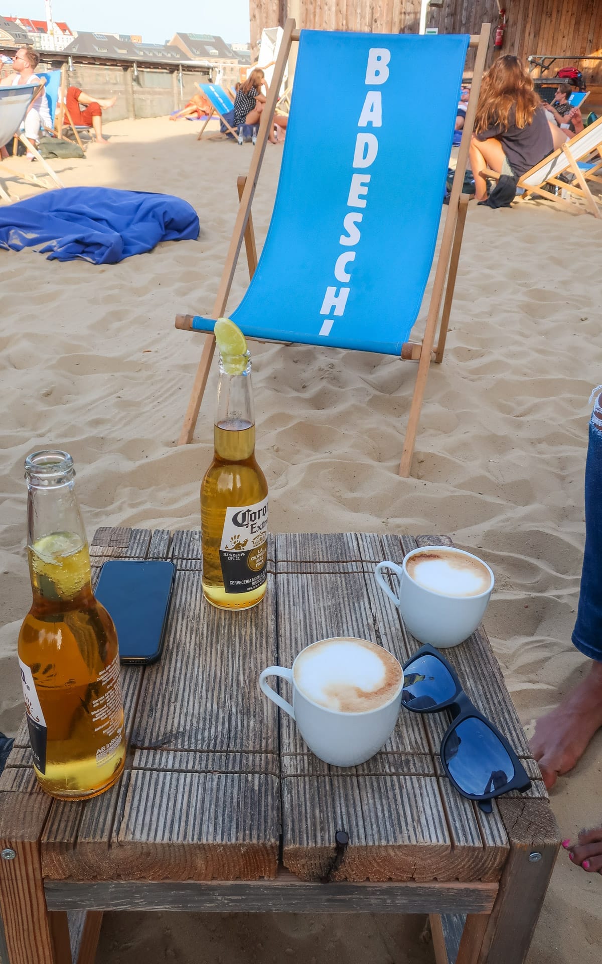 A wooden table on sandy ground holds beers and cappuccinos, with a blue deck chair at Badeschiff, Berlin’s beach club hotspot.