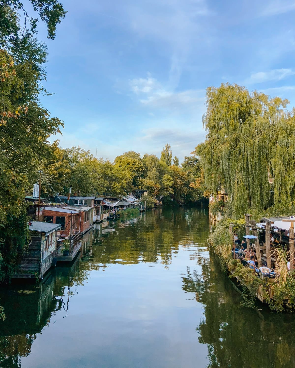 A peaceful canal in Berlin with floating houseboats, lush green trees, and an outdoor riverside cafe reflecting in the water.