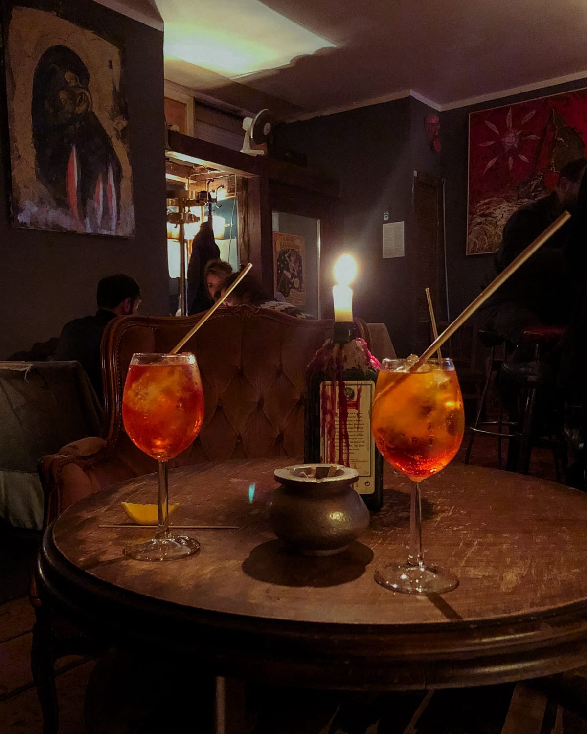 A cozy Berlin bar with dim lighting, eclectic art, and two Aperol Spritz cocktails on a rustic wooden table with a lit candle in a Jagermeister bottle.