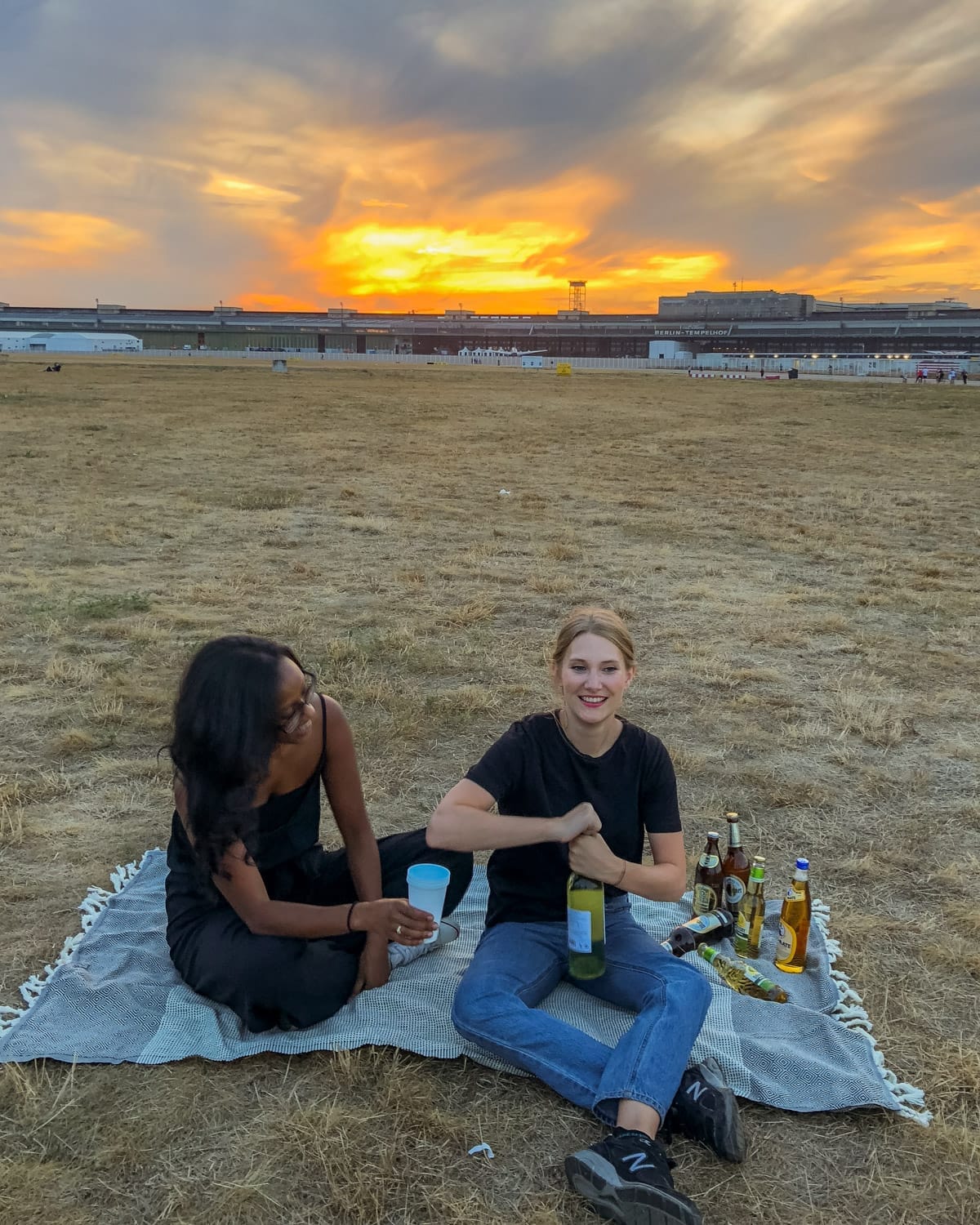 Two friends enjoying a sunset picnic at Tempelhofer Feld in Berlin, sitting on a blanket with bottles of wine and beer.