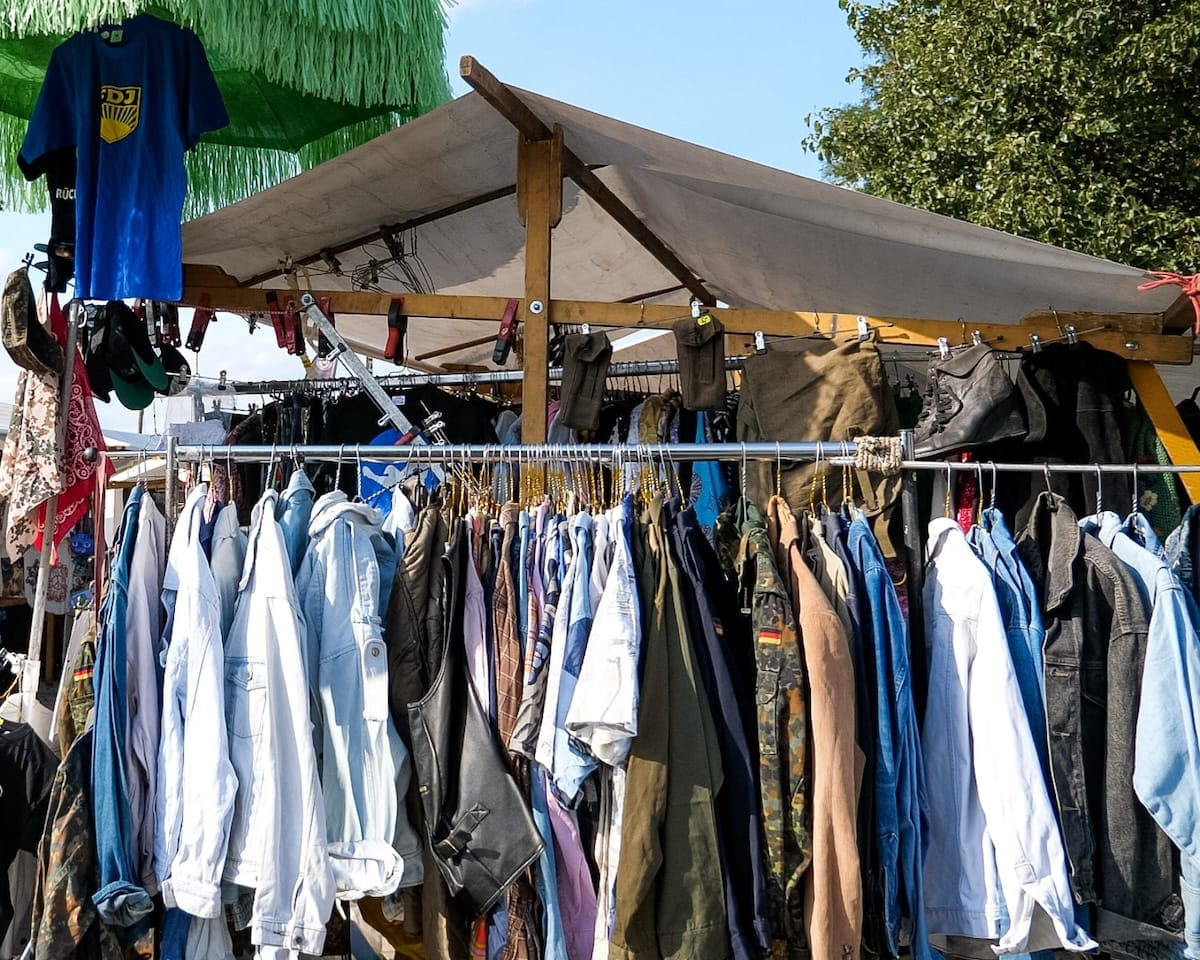 A stall at Mauerpark Market displays racks of secondhand denim, jackets, and military-style clothing under a canvas tent.